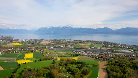 Vista-Aérea-De-Los-Campos-Rurales-Cerca-Del-Patio-De-Clasificación-De-Denges-Con-El-Lago-Leman-Y-Los-Alpes-De-Montaña-En-El-Fondo-En-Vaud,-Suiza
