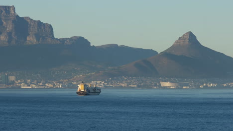Cargo-Boat-Floating-In-The-Sea-With-Table-Mountain-And-Lion's-Head-In-The-Background-In-Cape-Town,-South-Africa