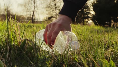 Picking-up-empty-plastic-water-bottle-lying-on-grass-park,-female-hand-grabs-rubbish,-golden-hour,-static-closeup