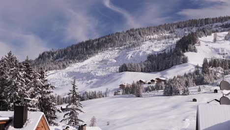 Fir-Trees-and-Roofs-of-Snowy-Houses-in-the-foreground-and-Ski-Slopes-and-Ski-Lifts-in-the-Italian-Alps-Mountains-Full-of-Snow-after-a-Heavy-Snowfall