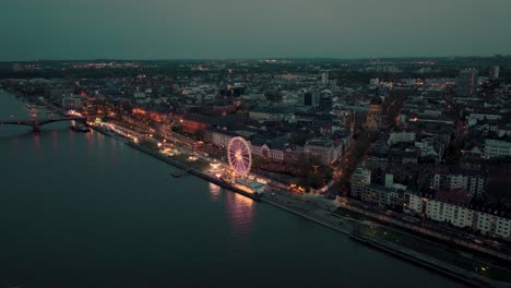 Drone-aerial-shot-wide-of-Mainz-coming-closer-showing-a-summer-night-wine-festival-at-the-Rhine-river-bank-with-lots-of-lights-under-the-sunset-with-the-Christus-church-and-the-cathedral