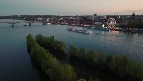 Sonnenuntergang-Drohnenflug-Von-Mainz-über-Den-Rhein-überquerung-Eines-Schiffes-Mit-Weinfest-Am-Ufer-Und-Sommerabendhimmel-Im-Rücken