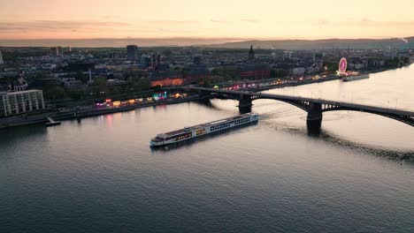 Sunset-drone-flight-over-Mainz-at-the-Rhine-river-with-a-boat-and-the-main-city-bridge-in-front-of-orange-sky-with-a-summer-wine-festival-in-the-back