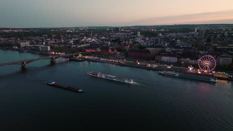 Drone-aerial-shot-of-Mainz-two-boats-crossing-in-front-of-a-summer-wine-festival-at-the-Rhine-river-bank-under-orange-sunset-sky