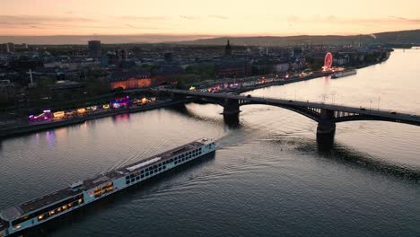 Sunset-drone-flight-of-Mainz-over-the-Rhine-river-crossing-a-boat-and-the-main-city-bridge-in-front-of-orange-sky