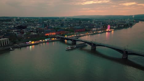 Aerial-Drone-Shot-of-Mainz-with-a-crossing-boat-and-the-main-Rhine-river-bridge-under-a-orange-summer-sunset-with-lots-of-reflections-on-the-water-and-a-wine-festival-in-the-back