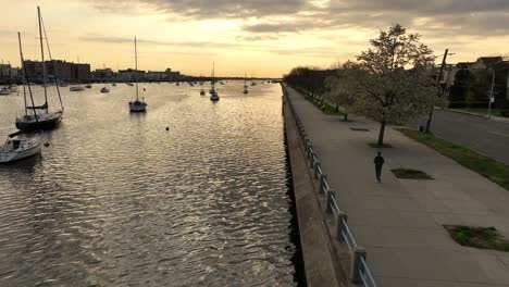 An-aerial-shot-over-Sheepshead-Bay-during-a-golden-sunrise-with-boats-anchored-and-a-jogger-running-on-the-paved-walkway-along-the-water