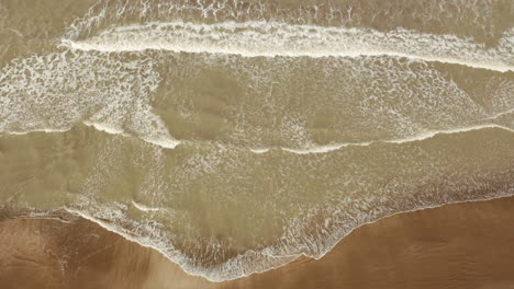 Aerial-view-of-small-sea-waves-advancing-towards-the-golden-sandy-coast-at-sunset