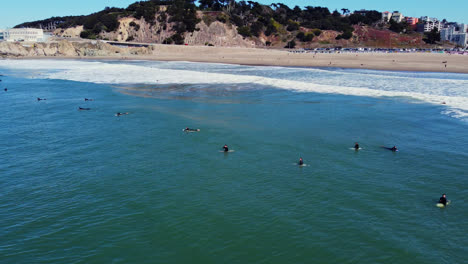 Surfistas-Acostados-Y-Sentados-En-Tablas-De-Surf-Flotando-En-El-Mar-En-Ocean-Beach-Con-Cliff-House-En-San-Francisco,-California