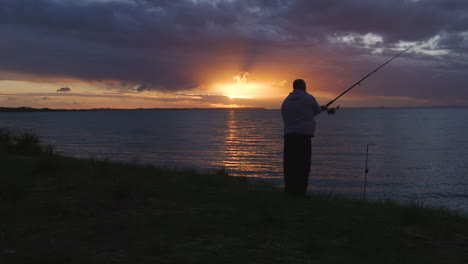 Ein-Einsamer-Fischer,-Der-Seine-Linie-Von-Den-Ufern-Des-Ruhigen-Wassers-Mit-Einem-Orangefarbenen-Sonnenuntergang-Am-Abend-Auswirft