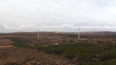 Approaching-rotating-wind-turbines-at-Lindesnes-windpark-in-southern-Norway---Two-turbines-operated-by-Asko-Fornybar---Aerial-in-flat-landscape-and-cloudy-background