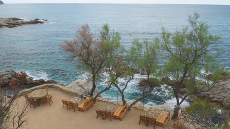 Cala-Banys-In-Lloret-De-Mar-Blick-Auf-Den-Felsstrand-Mittelmeer-Transparentes-Türkisblaues-Wasser-Spektakuläres-Restaurant-Direkt-Am-Meer-Mit-Chill-out-stühlen-Und-Direktem-Blick-Auf-Die-Klippen