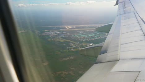 Airplane-Flying-Over-Leaving-Cancun-International-Airport-Seen-From-Passenger-Window-With-Airplane-Wing-In-The-Foreground