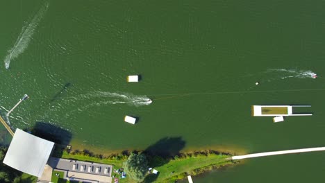 Birds-eye-view-drone-shot-of-a-waterski-system---drone-is-ascending-over-the-lake-with-waterskiers-and-wakeboarders-passing-by