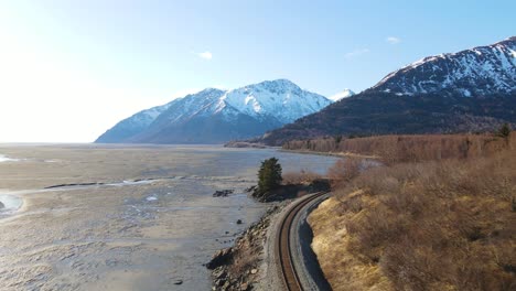 4K-drone-flying-over-railroad-tracks-along-the-cost-of-Alaska-with-snow-covered-mountains-in-the-background-on-a-sunny-day
