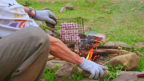 Close-up-of-a-person-moving-a-barbecue-grill-with-food-over-a-campfire-for-cooking