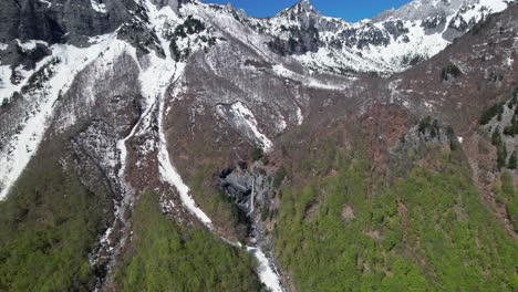 Waterfall-on-the-slope-of-the-alpine-mountain-covered-with-white-snow-in-Valbona-valley,-Albania