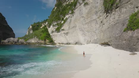 Woman-in-black-bathing-suit-running-on-white-sand-Diamond-Beach-in-Nusa-Penida,-steep-rock-cliffs,-tropical-paradise,-aerial