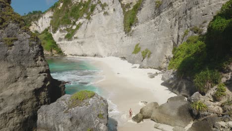 Diamond-beach-in-Nusa-Penida-with-steep-cliffs-and-woman-running-between-rocks,-aerial