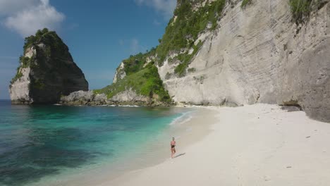 Woman-in-bikini-running-on-white-sand-beach-with-steep-cliff-on-tropical-island