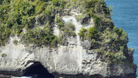 Rising-in-front-of-large-sea-rock-pinnacle-with-cave,-revealing-distant-fishing-boat,-Nusa-Penida,-aerial