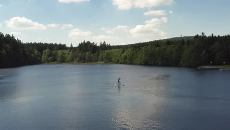 A-lone-person-paddling-a-stand-up-paddleboard-across-a-calm-pond-on-vacation,-aerial-orbit