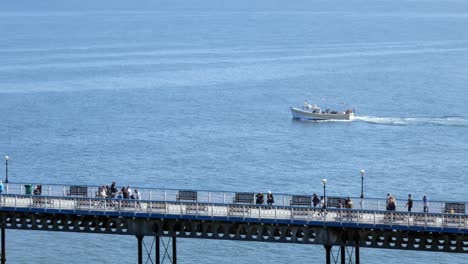 Sightseeing-boat-cruise-passing-tourists-walking-along-Llandudno-pier-seafront-boardwalk