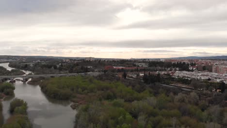 Rising-drone-over-river-and-industrial-area-with-park-on-cloudy-day