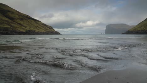 Close-up-of-flowing-water-of-Sea-reaching-shore-of-Tjörnuvik-beach---Risin-og-Kellingin-sea-stacks-in-background-during-cloudy-day-on-Streymoy-Island,-Faroe-Islands