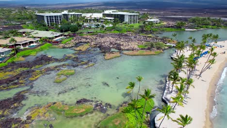 Tidal-pools-and-palm-trees-along-a-narrow-sandy-beach-with-hotels-and-resorts-in-the-background---descending-aerial-view