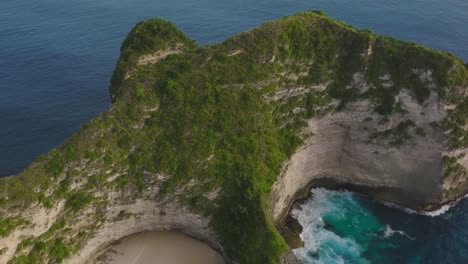 Steep-limestone-cliff-covered-in-green-vegetation-surrounded-by-blue-sea-water,-KelingKing-Beach,-aerial