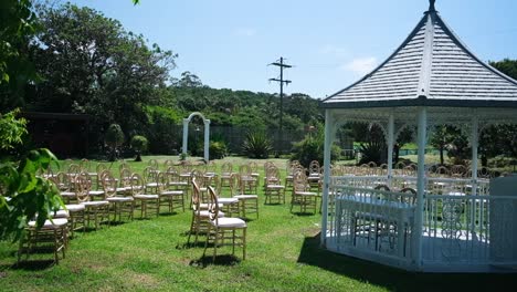 Stunning-Wedding-Gazebo-for-an-outdoor-wedding-under-the-trees,-accompanied-by-beautiful-sunny-weather-and-a-lovely-setup-of-gold-chairs-for-guests
