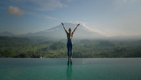 Attractive-blond-European-woman-on-infinity-pool-edge-stretching-into-upward-salute-yoga-pose,-Mount-Agung,-sunrise