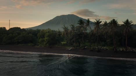 Traditional-outrigger-canoe-boat-arriving-at-tropical-shore-early-morning,-mount-Agung-in-background,-aerial
