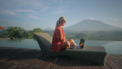 Mujer-Milenaria-Trabajadora-Escribiendo-En-Una-Laptop-Tendida-Hacia-Atrás-En-Un-Sillón-En-La-Piscina-Infinita,-Increíble-Paisaje-De-Bali