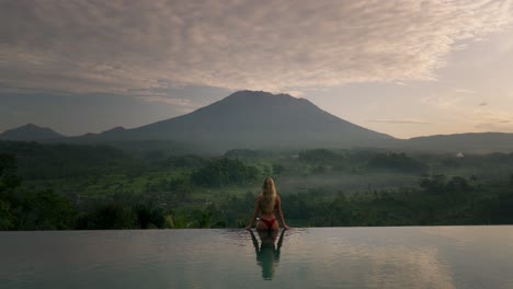 Blond-caucasian-woman-in-bikini-sitting-on-infinity-pool-edge-overlooking-magical-Mount-Agung-at-dawn