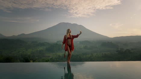 Fit-attractive-blond-woman-in-red-bikini-standing-on-infinity-pool-edge-taking-selfie,-Mount-Agung-volcano-in-background,-sunrise