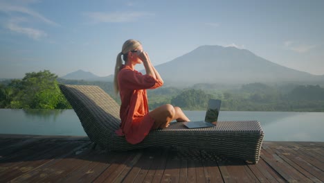 Confident-blond-business-woman-in-bathrobe-working-on-laptop-whilst-enjoying-view-of-Mount-Agung-at-pool