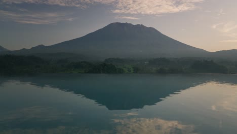 Volcano-Mount-Agung-during-hazy-sunrise,-infinity-pool-reflection-on-water-surface,-tropical-landscape