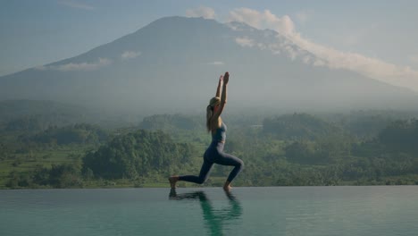 Fit-woman-in-sportswear-standing-in-Warrior-Position-stretching-arms-above-head,-Mount-Agung-in-background,-bright-morning-sunlight