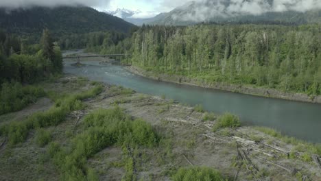 Drone-footage-of-beautiful-Pacific-Northwest-river,-misty-forest-with-bridge-and-mountains-in-background