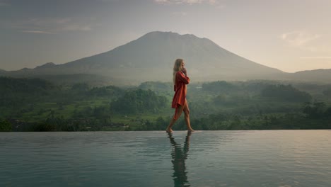 Blond-business-woman-in-silk-bathrobe-walking-on-infinity-pool-edge-while-on-phone-call,-Mount-Agung-in-background
