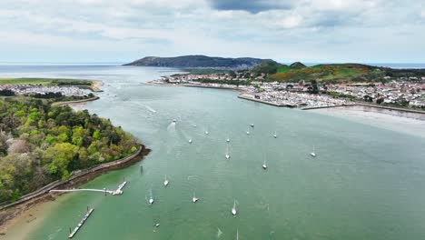 Several-sailboats-anchor-in-the-fast-flowing-Conwy-River-with-the-coastal-town-of-Deganwy-amongst-the-green-hills-of-Welsh-nature-on-a-cloudy-day