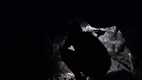Female-explorer-crouching-in-dark-Kaumana-Lava-Cave,-lighting-cavern-with-head-torch