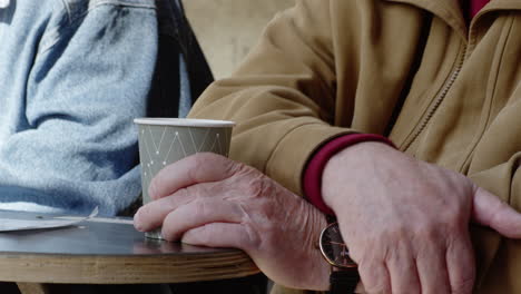 ältere-Freunde-Sitzen-Am-Parkbanktisch-Trinken-Kaffeetasse-Café-Reden-Diskutieren-Diskutieren-Gespräch-Erinnerungen-Nahaufnahme-Hände-Hand-Gespräch-Unterhalten-Sich-Hinsetzen-Geduld-Warten-Restaurant
