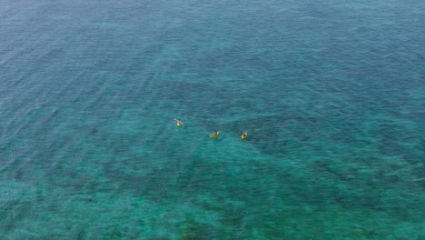 Three-people-in-yellow-kayaks-paddling-in-shallow-blue-tropical-water,-aerial