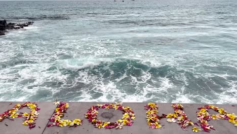 Aloha-spelled-out-in-colorful-flower-petals-welcoming-guests-as-the-lookout-over-waves-crashing-into-the-Hawaiian-beach