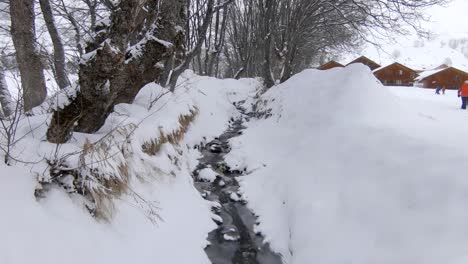 A-small,-partly-frozen-stream-flowing-down-a-snowy-winter-landscape-with-trees-and-some-chalets-in-the-background