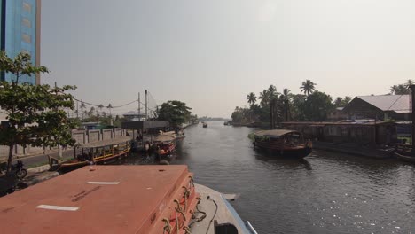 Pov-boat-cruise-along-waterway,-wooden-boats-on-shore,-Kerala-Local-lifestyle