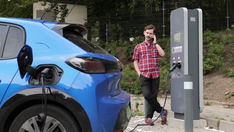 Young-man-chat-on-mobile-phone-while-electric-car-is-charging-on-outdoor-station